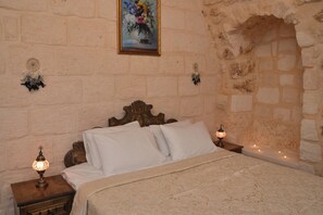 Un dormitorio con pared de piedra, cabecera de madera, cama con ropa blanca, mesita de noche con lámpara y un cuadro de flores en la pared.
