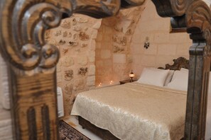 Un dormitorio con pared de piedra, cabecera de madera, cama con colcha blanca y una vela encendida en la mesita de noche.