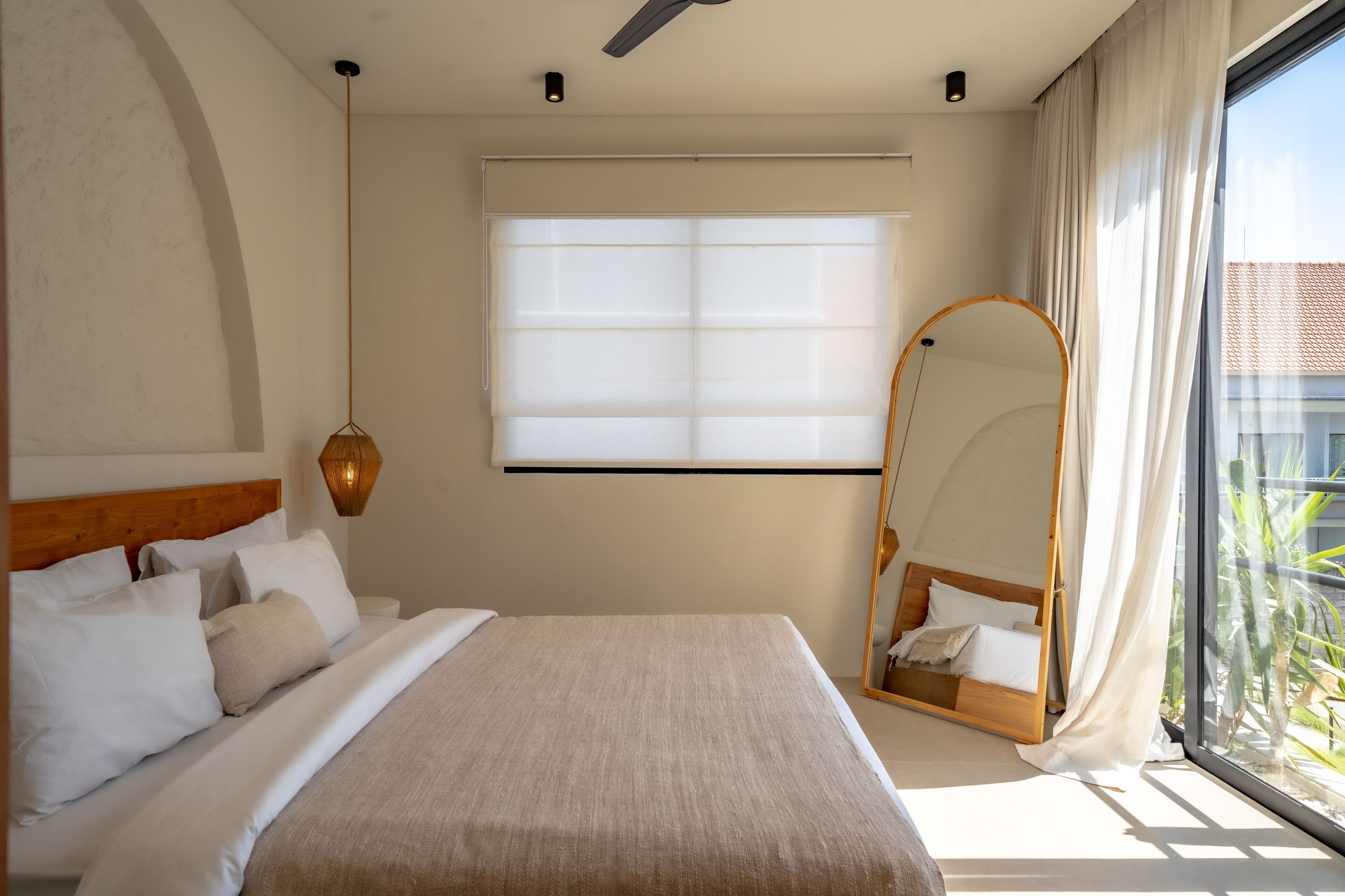 A bedroom with a large bed, a ceiling fan, a window with sheer curtains, a hanging lamp, and a large mirror.
