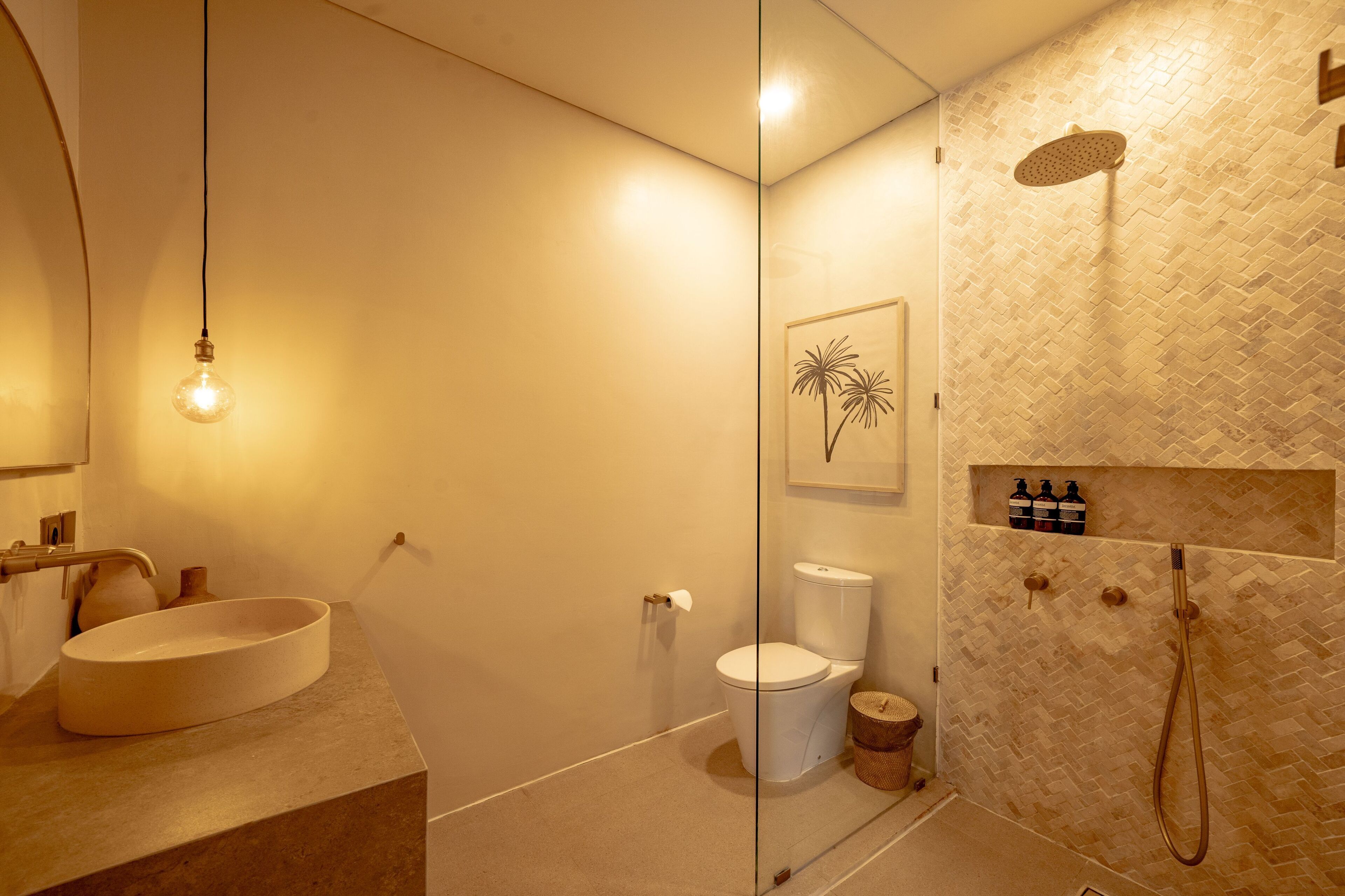 Un baño moderno con un lavamanos ovalado grande, un inodoro, ducha con cabezal de ducha manual y un estante de pared con artículos de aseo.