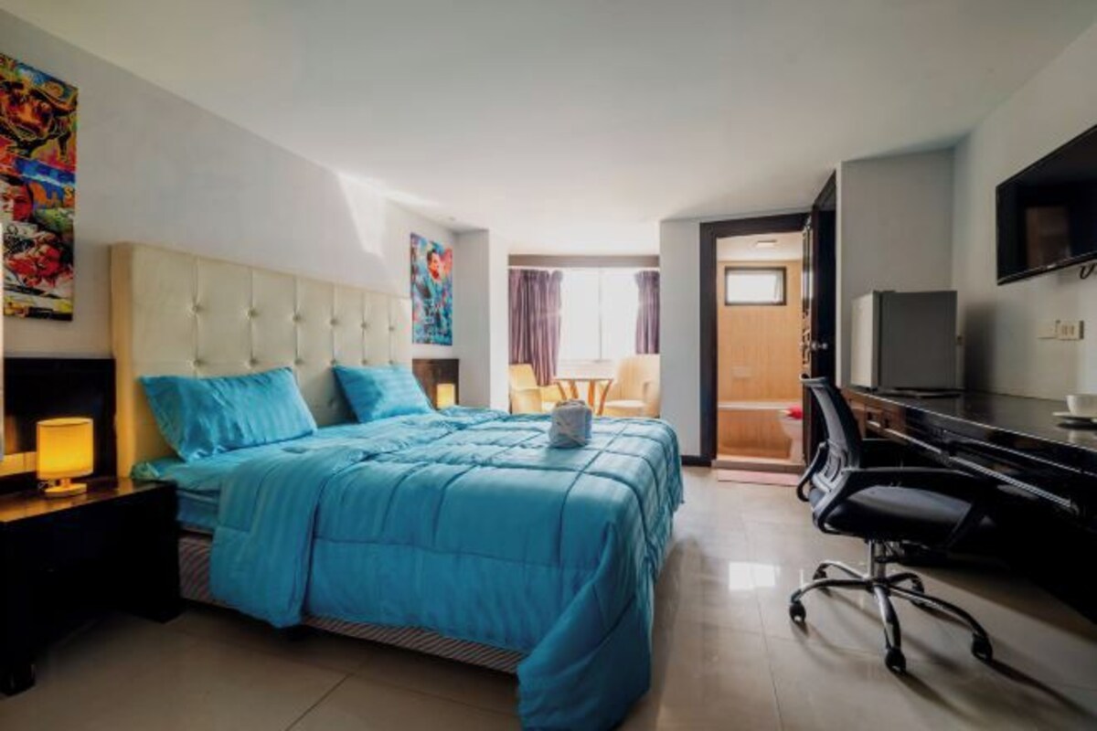 A hotel room with a bed, a desk, a chair, a TV, and artwork on the walls.