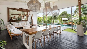 A modern outdoor dining area with a wooden table and chairs, surrounded by lush greenery and a pool. - Umalas 5BR Bohemian Escape • Rice Fields + Yoga & Spa w/ 12min To Beach (Badung)