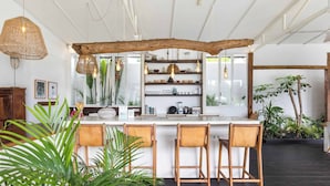 A modern kitchen with a central island, wooden chairs, hanging pendant lights, and a rustic wooden beam above. - Umalas 5BR Bohemian Escape • Rice Fields + Yoga & Spa w/ 12min To Beach (Badung)