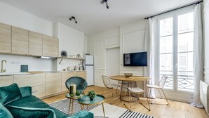 Ein modernes Wohnzimmer mit einem grünen Sofa, einem Esstisch mit Stühlen, einem Fernseher und einer Küchenzeile mit Schränken und Geräten.