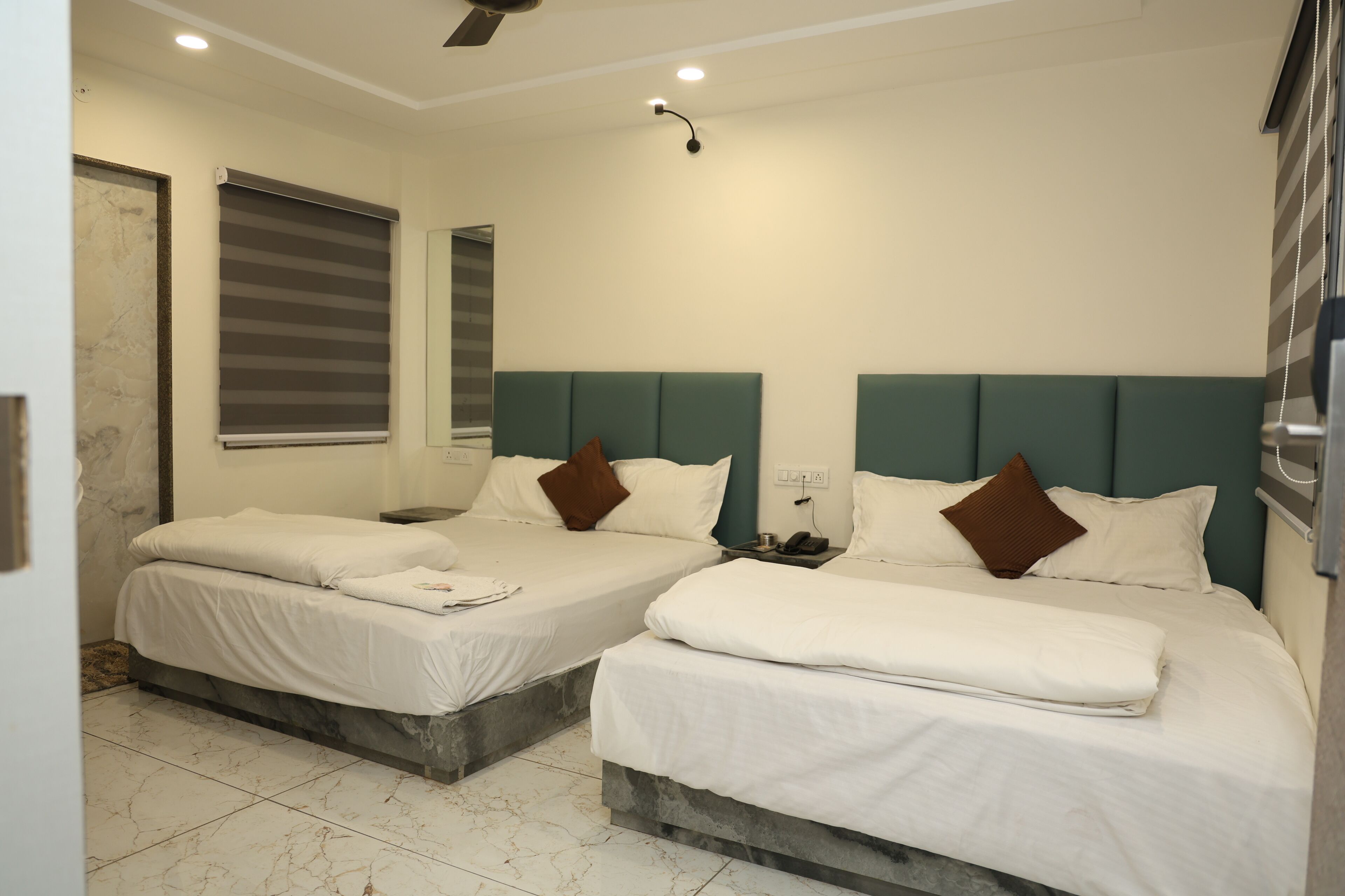 Habitación de hotel con dos camas, cada una con cabecero, ropa de cama blanca y almohada marrón. La habitación tiene piso de baldosas y ventilador de techo.