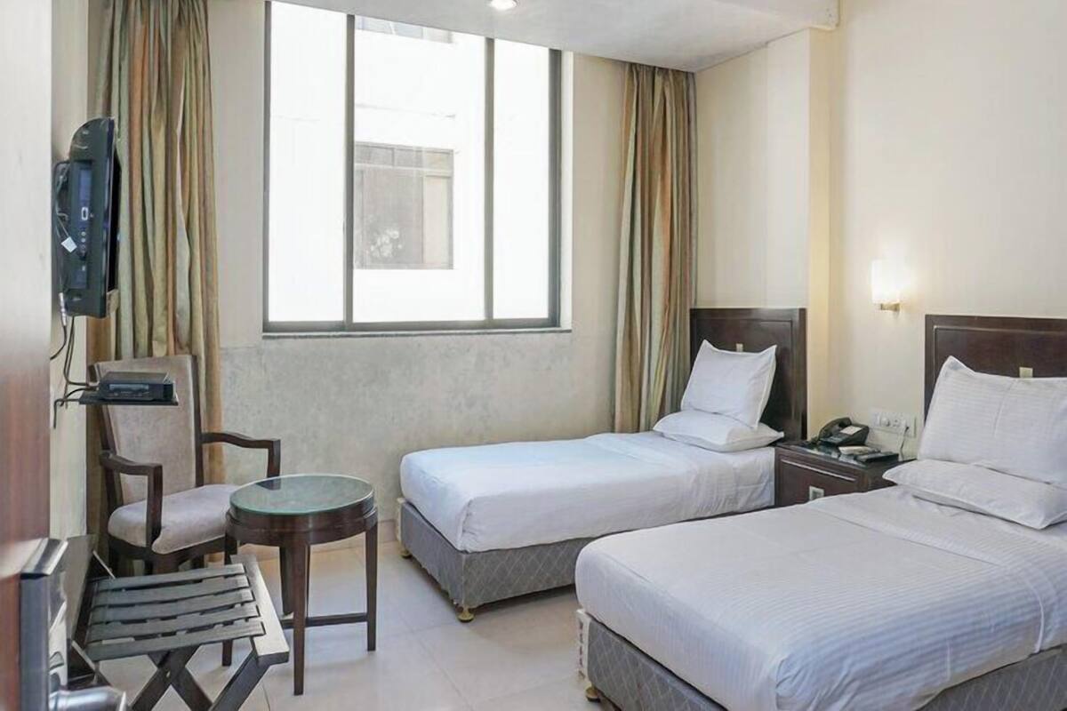 Ein Hotelzimmer mit zwei Einzelbetten, einem kleinen Tisch, einem Stuhl, einem Fernseher und einem Fenster mit Vorhängen.