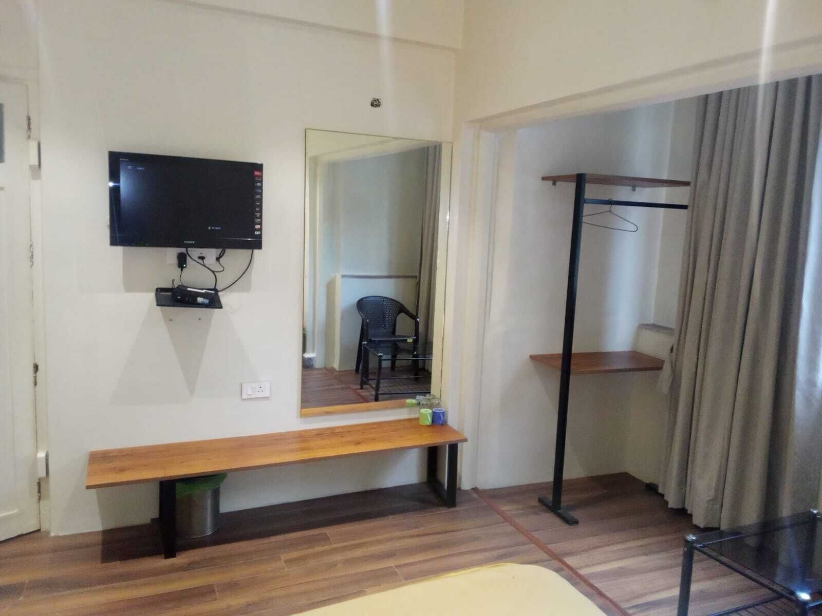 Ein Zimmer mit einer Holzbänkl, einem an der Wand montierten Fernseher, einem Spiegel, einem Sessel und einem Kleiderständer.