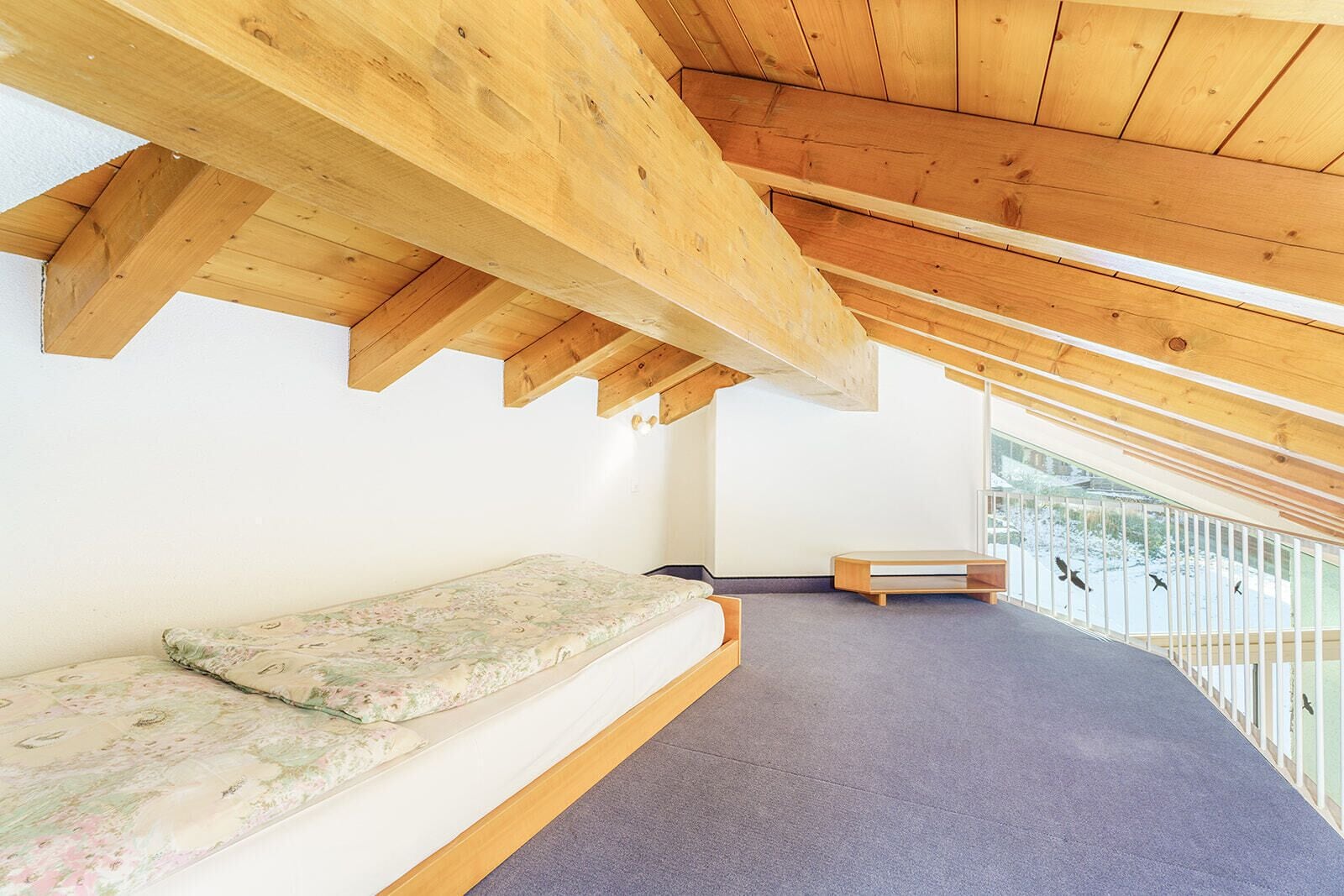 Una habitación con techos altos y vigas de madera, una cama con colchón floral, un banco y un balcón con barandilla.