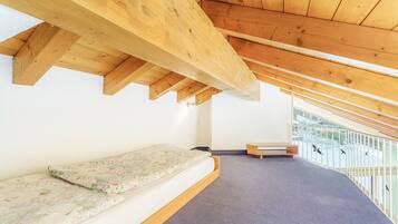 Una habitación con techos altos y vigas de madera, una cama con colchón floral, un banco y un balcón con barandilla.