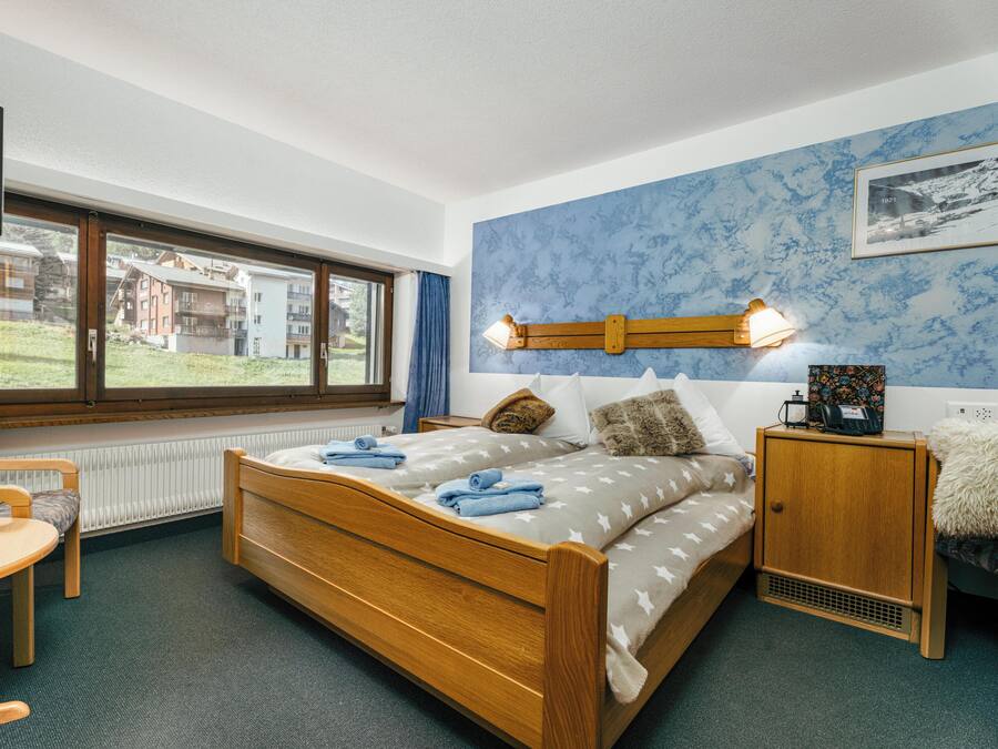Un dormitorio con cama de madera, una mesita de noche, una silla, una ventana con vistas a edificios y un cuadro en la pared.