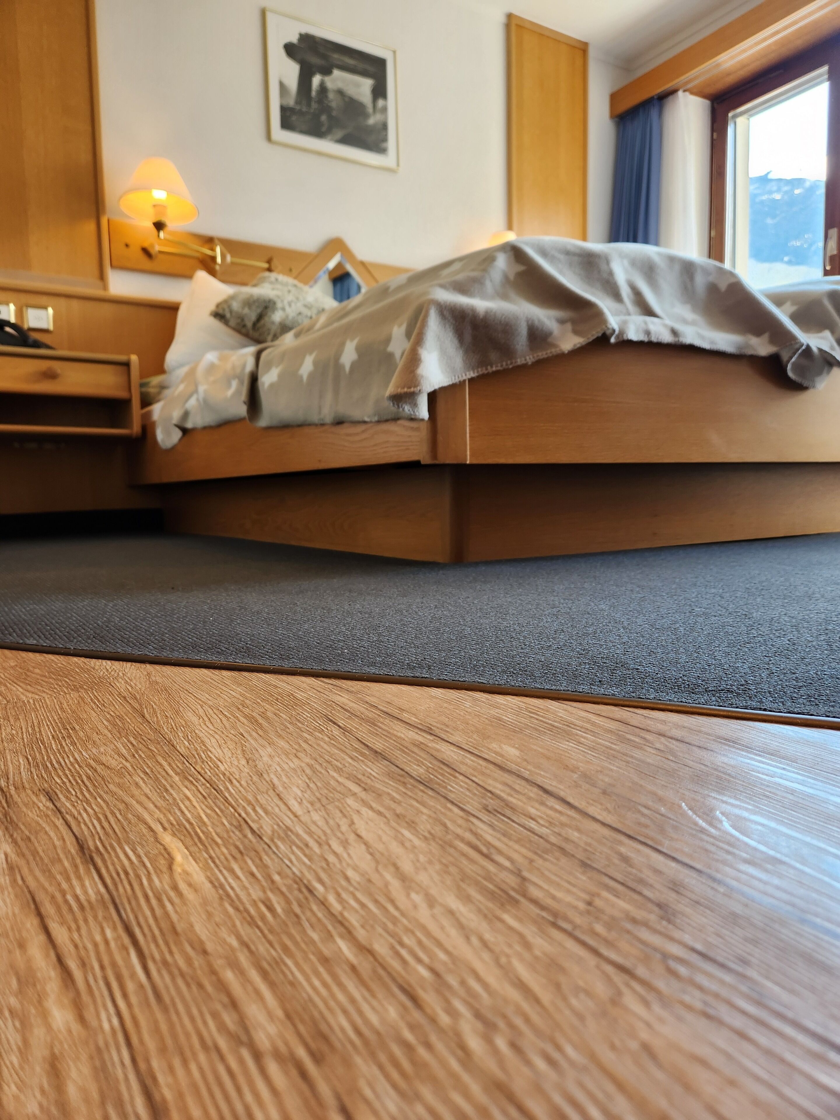 Una cama de madera con una manta gris, una mesita de noche con una lámpara y un cuadro enmarcado en la pared.