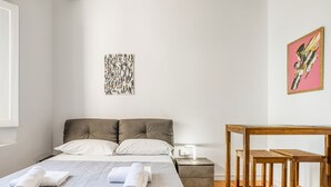 Een slaapkamer met een bed, twee kussens, een nachtkastje met een lamp, een houten nachtkastje en een kleine tafel met krukjes.