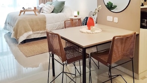 A modern hotel room with a bed, a dining table with chairs, a hanging light fixture, and a round mirror on the wall.
