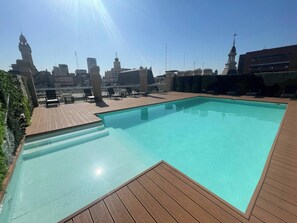A rooftop swimming pool with wooden deck and lounge chairs, surrounded by city buildings and clear skies. - Modern Studio at the Heart of San Telmo With Pool (Buenos Aires)