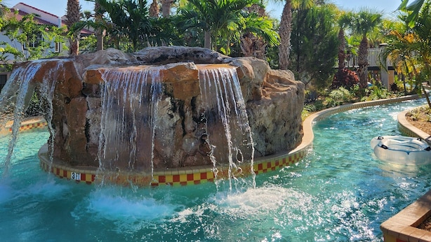 A pool with a waterfall feature surrounded by palm trees and a clear blue sky. - Stop Scrolling - FREE Water Park Access - Newly Renovated Family Resort Home (Kissimmee)