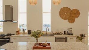 Una cocina moderna con una isla grande, dos lámparas colgantes, una estufa integrada y vistas al agua a través de las ventanas.
