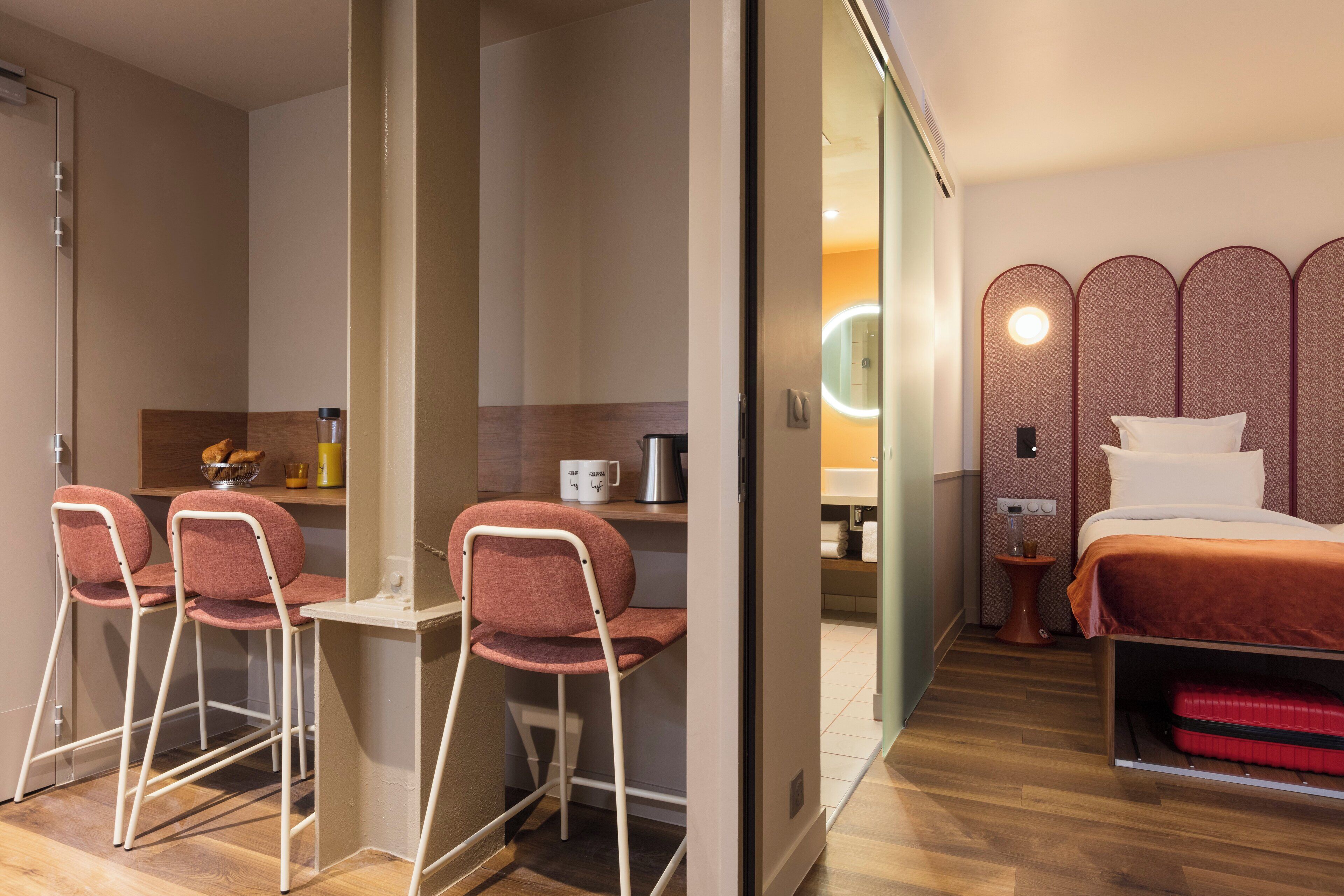 Habitación de hotel pequeña y funcional, con una cama, un escritorio con silla, un espejo y una mesita de noche.