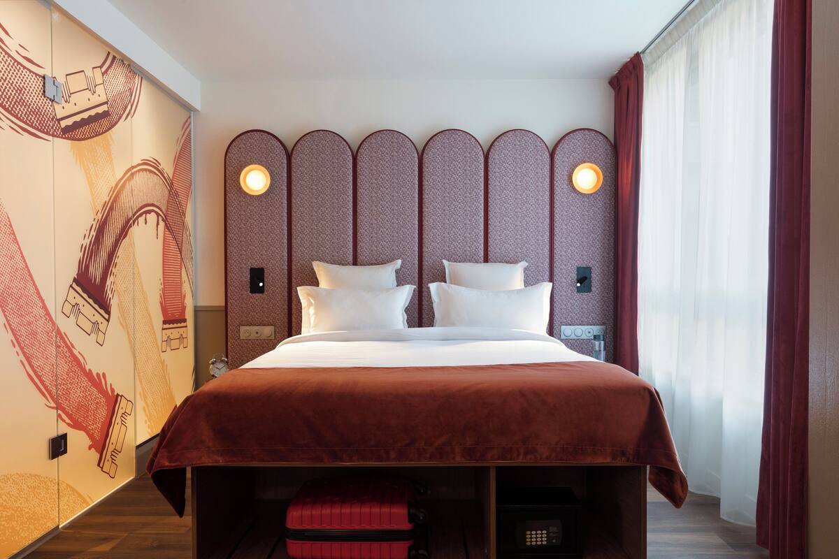A hotel room with a large bed, a headboard with lights, a red suitcase, and a wall with a hand-drawn design.