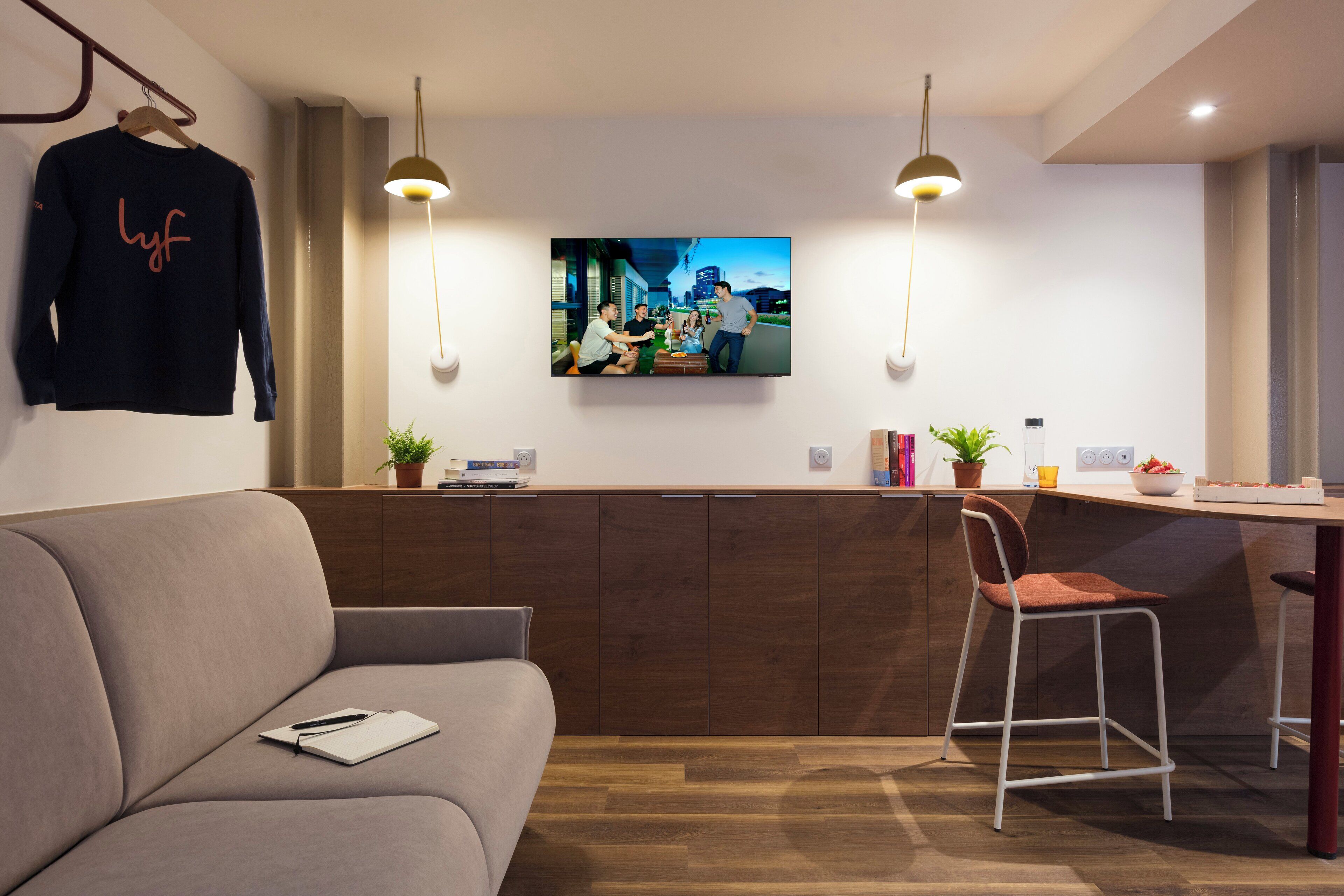 A modern living room with a sofa, a dining area with a bar, and a TV mounted on the wall displaying a cityscape.