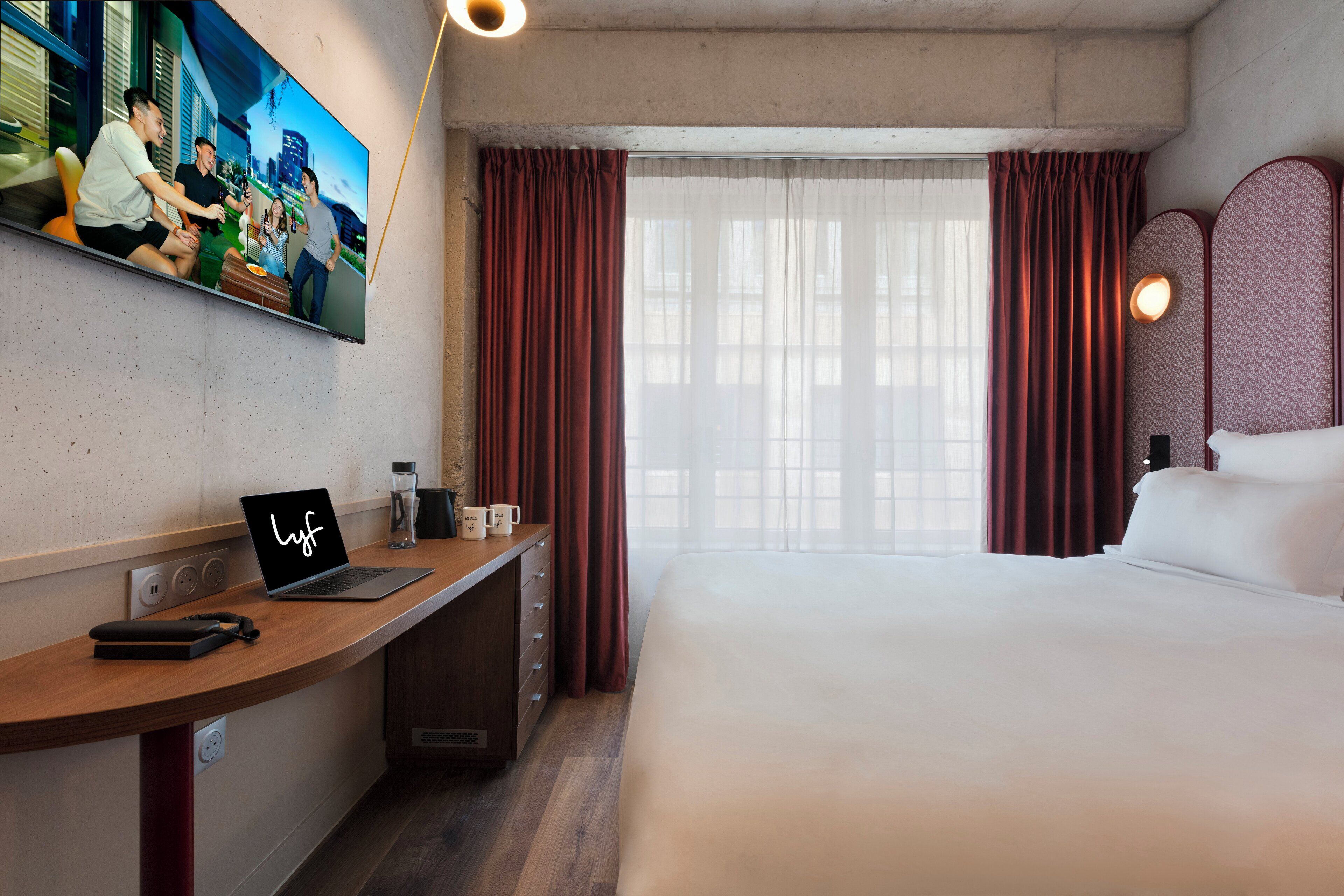 Habitación de hotel con cama, escritorio con ordenador portátil, televisión en la pared y ventana con cortinas.