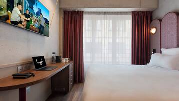 Habitación de hotel con cama, escritorio con ordenador portátil, televisión en la pared y ventana con cortinas.