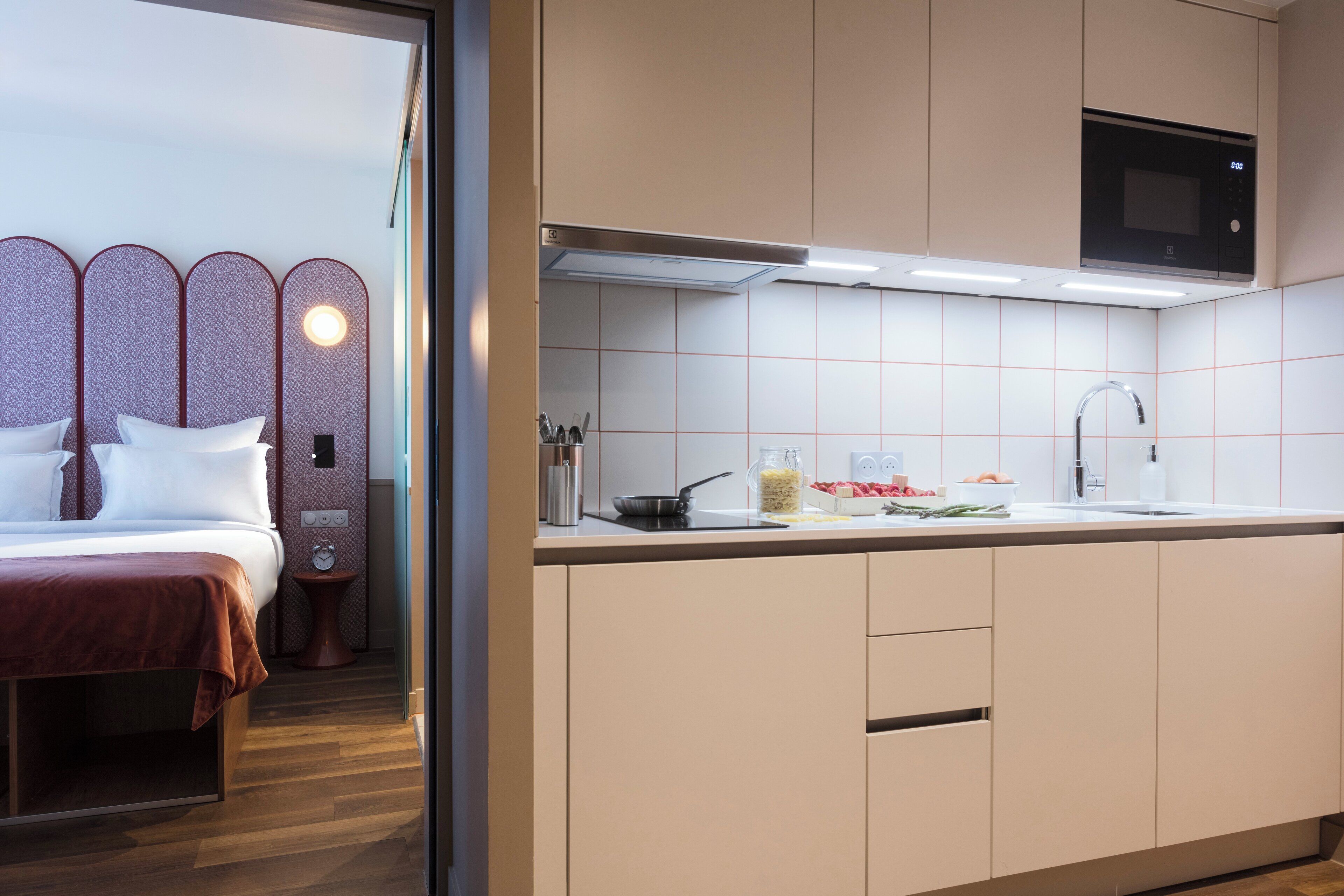Una habitación de hotel moderna con una cama, una mesita de noche y una kitchenette que incluye microondas, lavamanos y armarios.