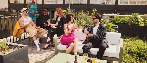 A group of people seated on outdoor furniture, with drinks on a table and an umbrella providing shade.