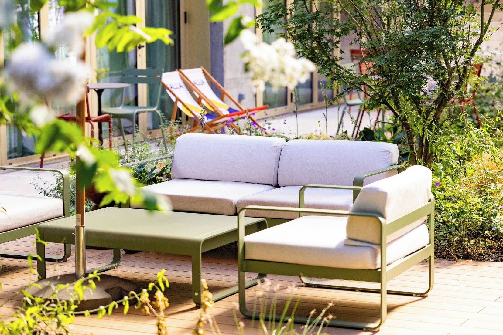 Un área de estar al aire libre con sofá y sillas, rodeada de plantas y vistas a un edificio.