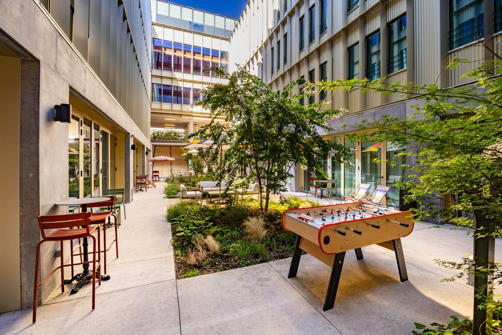 An outdoor seating area with tables and chairs, a foosball table, and a garden with plants and trees.