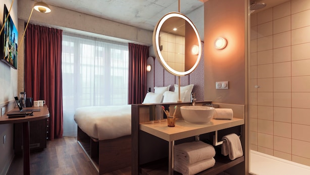 Une chambre d’hôtel avec un lit, un bureau avec un ordinateur portable, un miroir rond, un lavabo et une douche.