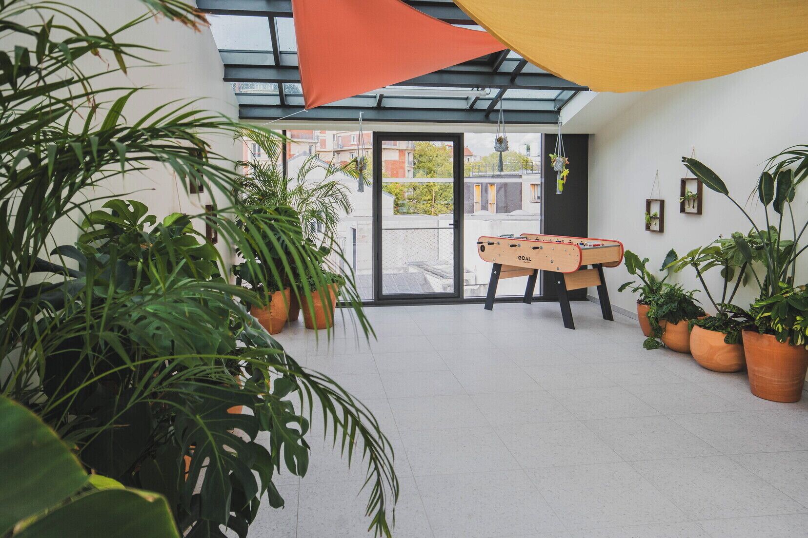 A room with a foosball table, potted plants, and a glass ceiling.