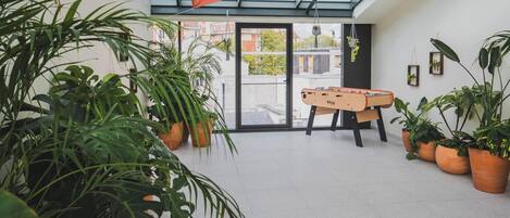 A room with a foosball table, potted plants, and a glass ceiling.
