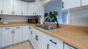 Una cocina moderna con armarios blancos, encimera de madera, pileta doble, microondas y una planta grande.