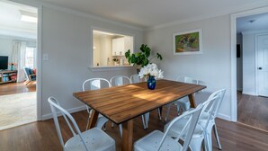 Un coin repas avec une table en bois et des chaises blanches, un vase rempli de fleurs et un tableau accroché au mur.