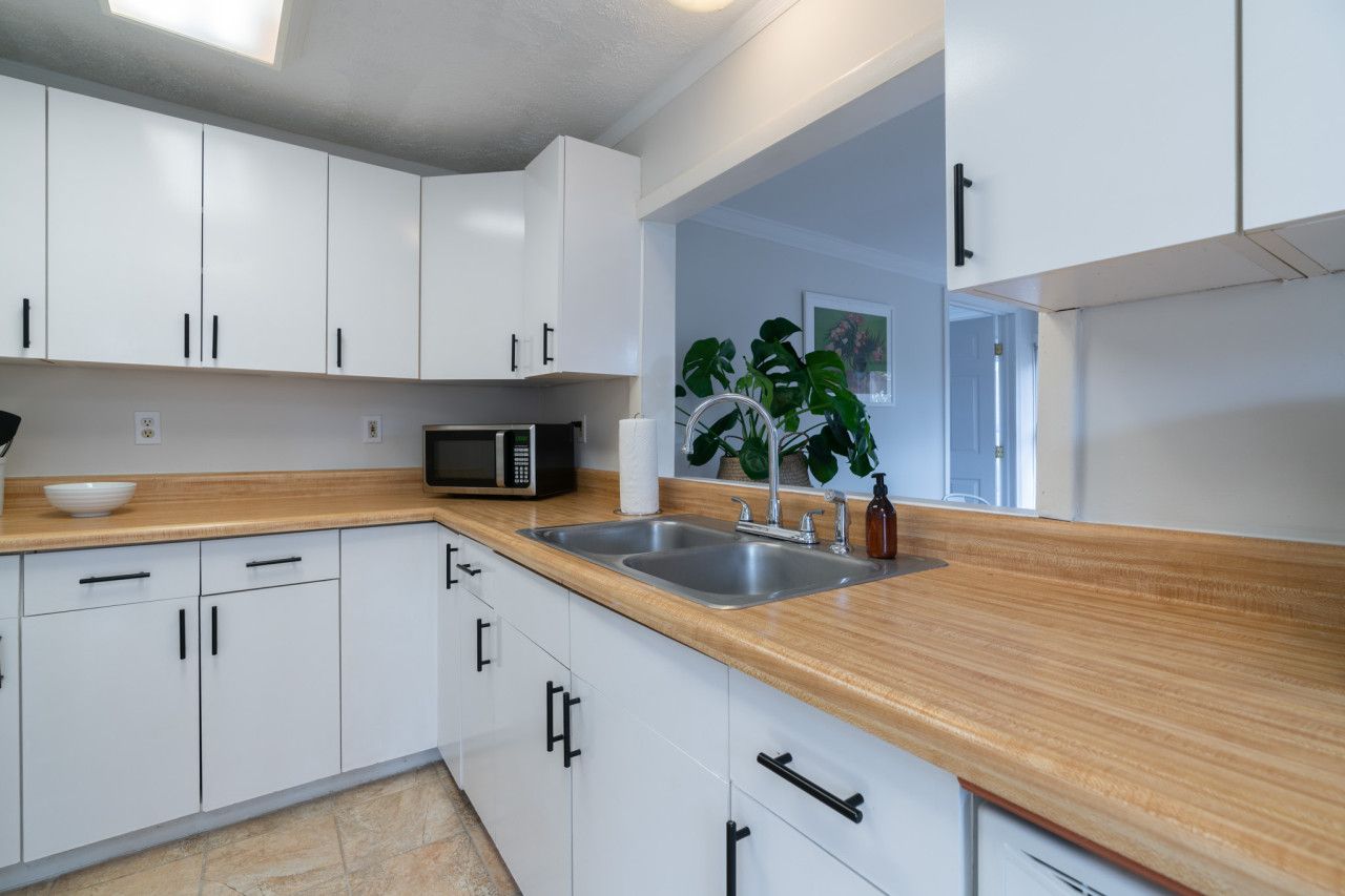 Une cuisine moderne avec des meubles blancs, un plan de travail en bois, un évier double, un four à micro-ondes et une grande plante.