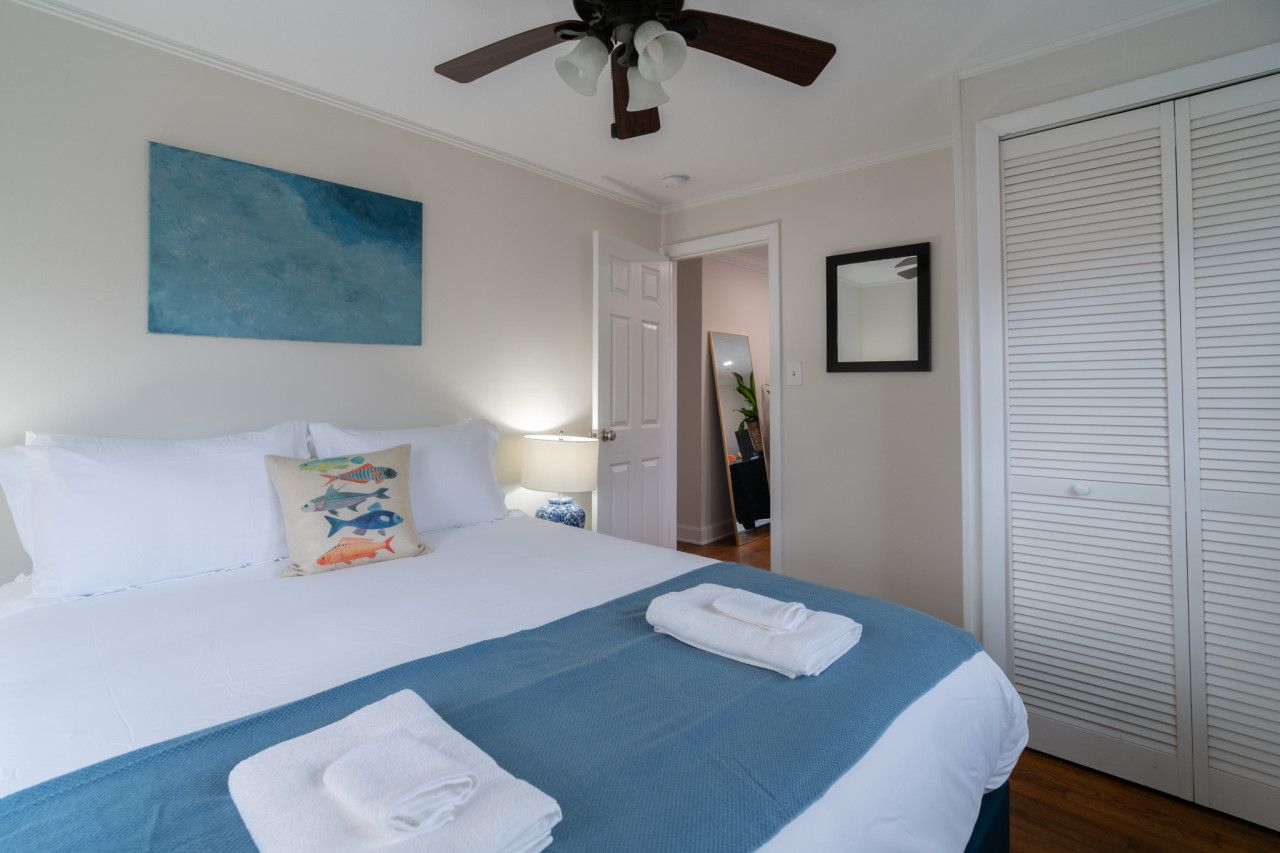 Ein ordentlich bezogenes Bett mit blauer und weißer Bettwäsche, ein Deckenventilator, ein blaues Bild an der Wand, ein Spiegel und ein weißer Kleiderschrank.