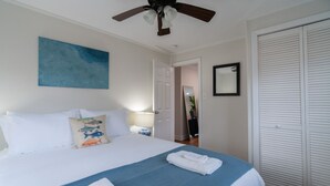 Un lit bien fait, avec une literie bleue et blanche, un ventilateur de plafond, un tableau bleu accroché au mur, un miroir et une armoire blanche.