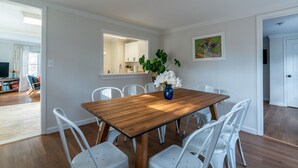 Un comedor con una mesa de madera y sillas blancas, un jarrón con flores y un cuadro en la pared.
