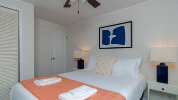 Ein Hotelzimmer mit einem Bett, zwei Nachttischen mit Lampen, einem Deckenventilator, einer Tür und einem gerahmten abstrakten Kunstwerk an der Wand.