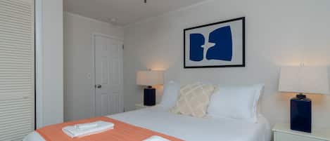 A hotel room with a bed, two bedside tables with lamps, a ceiling fan, a door, and a framed abstract artwork on the wall.