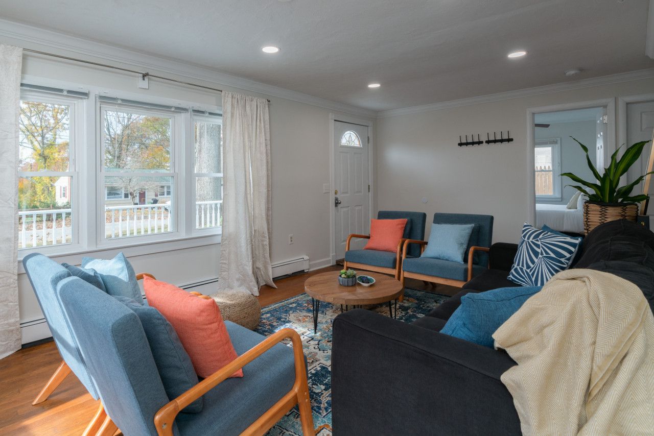 A living room with a large window, a black sofa, two blue armchairs, a wooden coffee table, and a patterned rug.