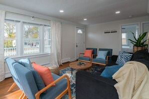 Een woonkamer met een groot raam, een zwarte bank, twee blauwe fauteuils, een houten salontafel en een kamerbreed tapijt met een patroon.