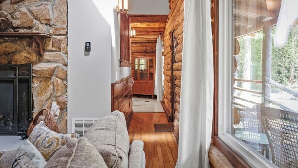 A cozy log cabin interior with a stone fireplace, wooden floors, and a view of the outdoors through a large window.