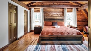 A bedroom with a wooden ceiling, a large bed, two windows with curtains, and a wooden floor.