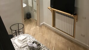 Et soveværelse med et fjernsyn på et træstel, en radiator under, en seng med et mønstret sengetæppe, og et lille bord med håndklæder.