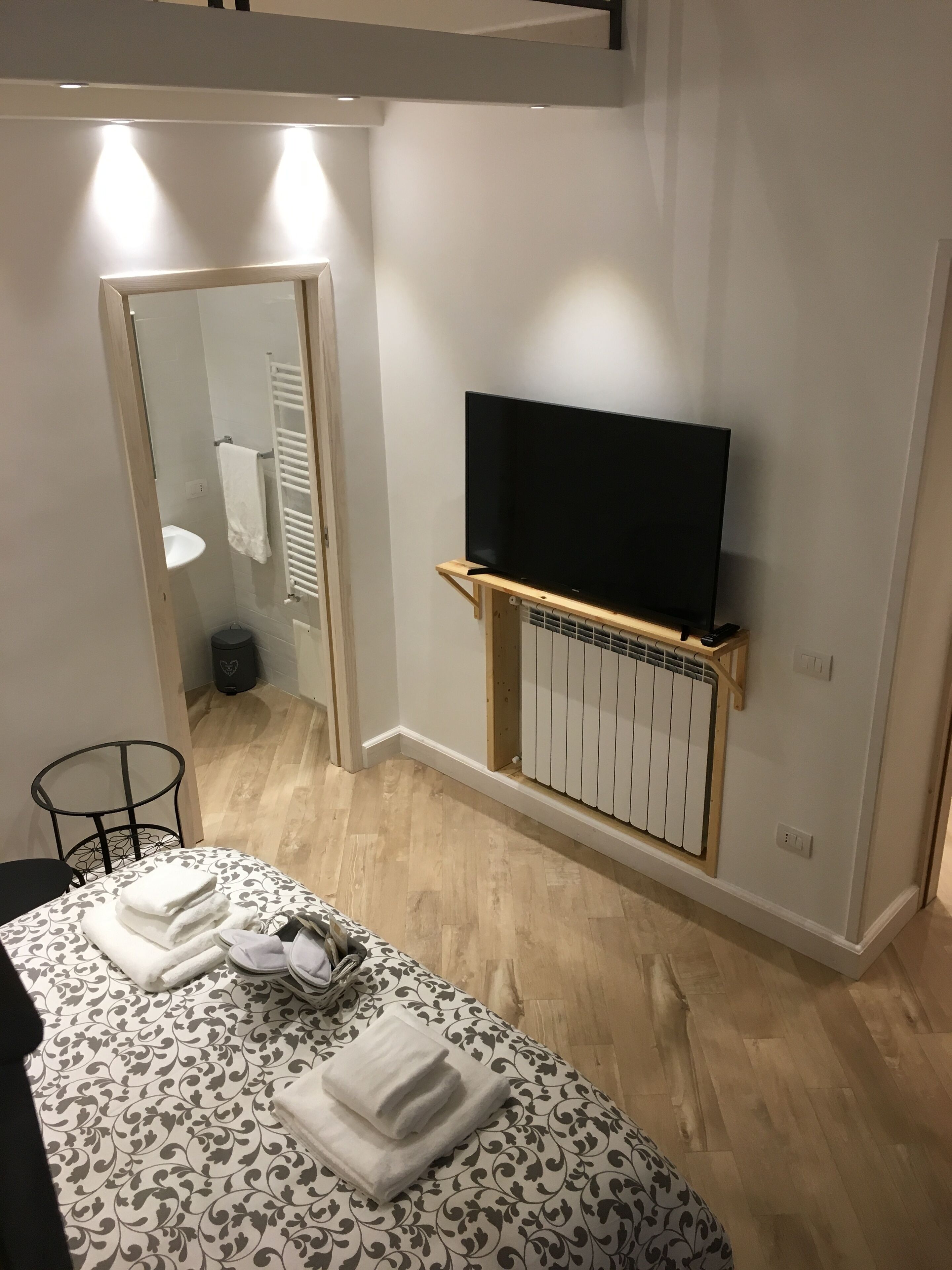 A bedroom with a bed, a TV mounted on a wooden shelf, a bathroom visible through an open door, and a small table with a chair.