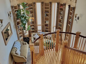 A spacious living area with a high ceiling, a wooden staircase, and a large bookshelf filled with books.