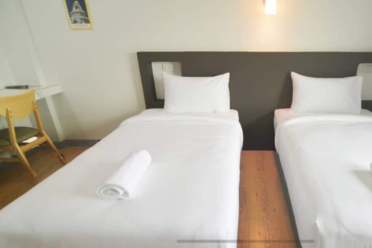 A hotel room with two single beds, each with a white duvet and a rolled towel. There is a wooden chair and a small table to the left.