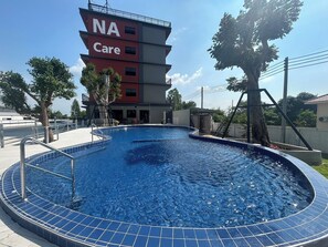 A swimming pool with a tiled deck, a building labeled "NA Care" in the background, and trees around the area. - Na Care Hotel (Chaiyaphum)