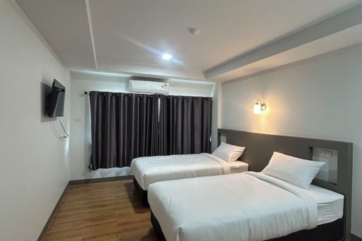 Habitación de hotel con dos camas, un televisor de pantalla plana montado en la pared, un equipo de aire acondicionado sobre las camas y una lámpara fijada a la pared.
